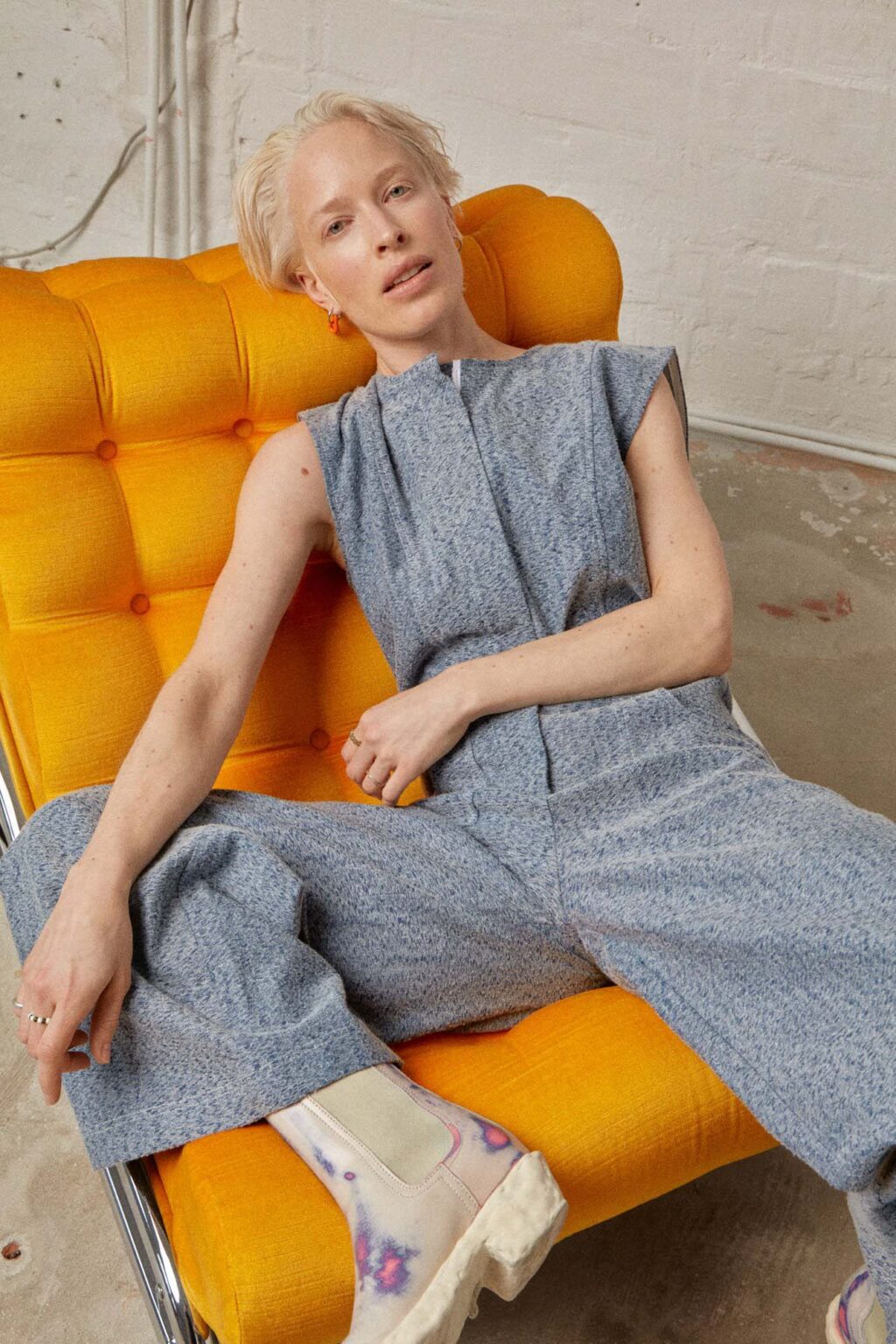 Woman in a blue/grey Jumpsuit sitting on a bright orange chair. Made by Atelier Dreisieben, Schneiderei Hamburg