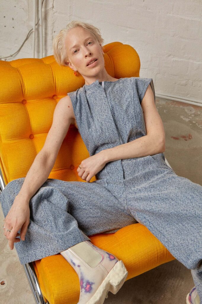 Woman in a blue/grey Jumpsuit sitting on a bright orange chair. Made by Atelier Dreisieben, Schneiderei Hamburg